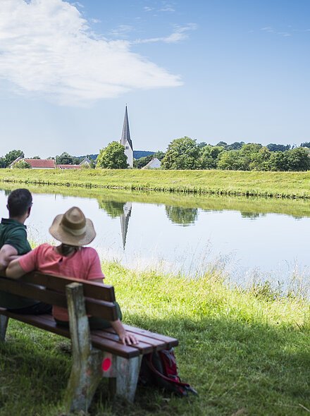 Poikam Kanal und Poikam Das Foto zeigt eine idyllische Uferlandschaft an einem ruhigen Fluss. In der Bildmitte erhebt sich der spitze Turm einer Kirche, der sich deutlich im Wasser spiegelt. Rechts und links der Kirche sind bäuerliche Gebäude mit roten Dächern zu erkennen, eingebettet in viel Grün. Zwei Menschen sitzen am Ufer auf einer Wiese. Der Himmel ist blau mit weißen, teils aufgelockerten Wolken. Die Szenerie wirkt friedlich und ländlich. Am Ufer der Donau sitzen zwei Wanderer.