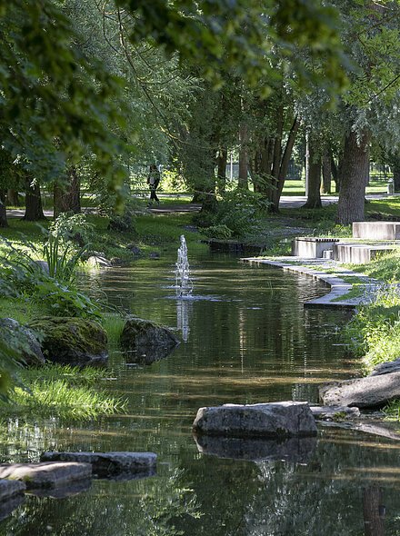 Markt Bad Abbach Kurpark Bachlauf Blick auf einen schmalen Bach mit Springbrunnen in einem Park mit Bäumen, Wiesen und Sitzgelegenheiten.