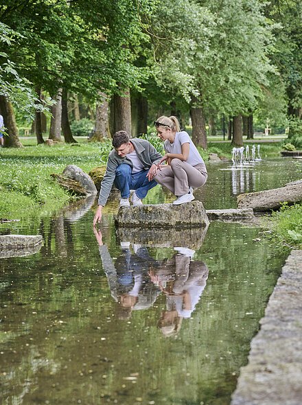 Bach im Kurpark Zwei Jugendliche sitzen auf einem Stein inmitten des Baches im Kurpark inmitten der grünen Idylle.