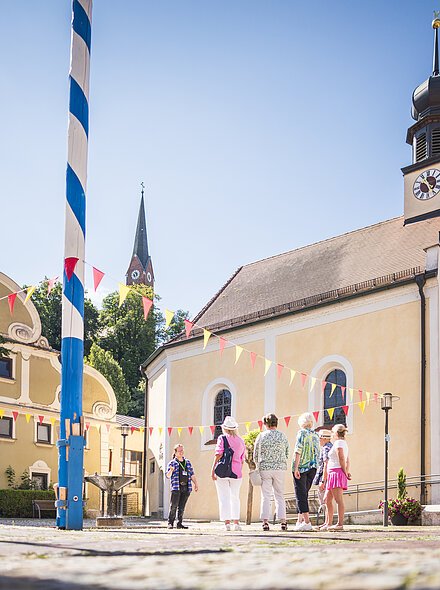 Gästeführung in der Marktmitte Vier erwachsene stehen in der Marktmitte neben der Marktkirche und folgen einem Gästeführer. Der Markt ist mit rot gelben Wimpeln dekoriert und links steht der blau-weiß gestreifte Maibaum steht unter blauem Himmel.